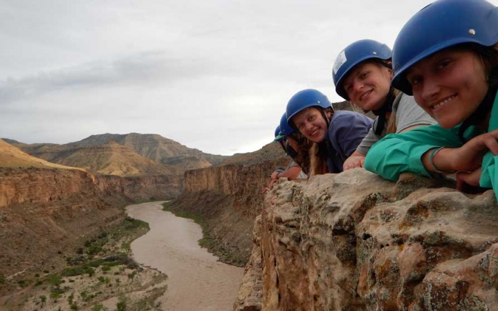 three people in helmets lay on a cliff overlooking the river below
